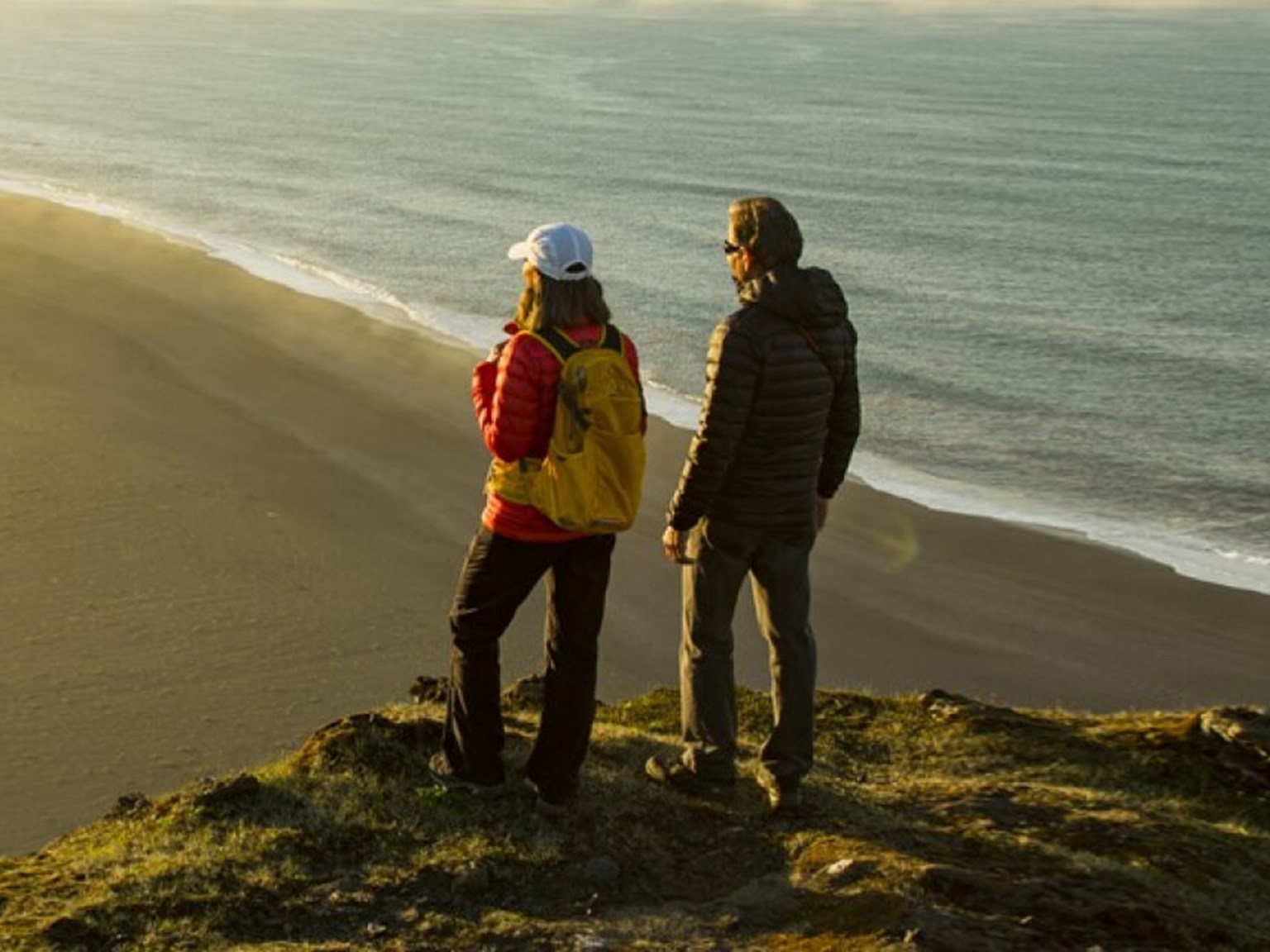 Active couple, stood on a cliff edge, overlooking a vast beach at sunset