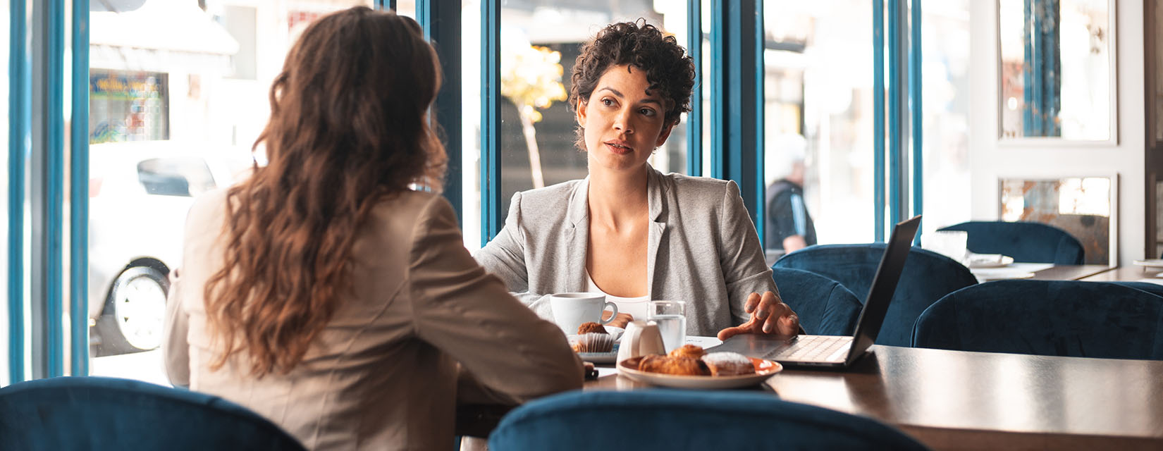 Two women talking business beside a laptop at a cafe over drinks and croissants