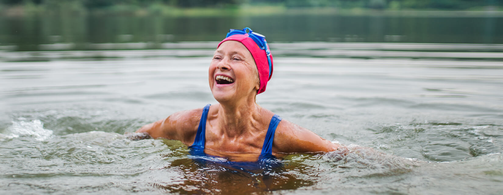 Happy older woman, wild swimming in a lake, wearing a pink swimming cap and blue goggles