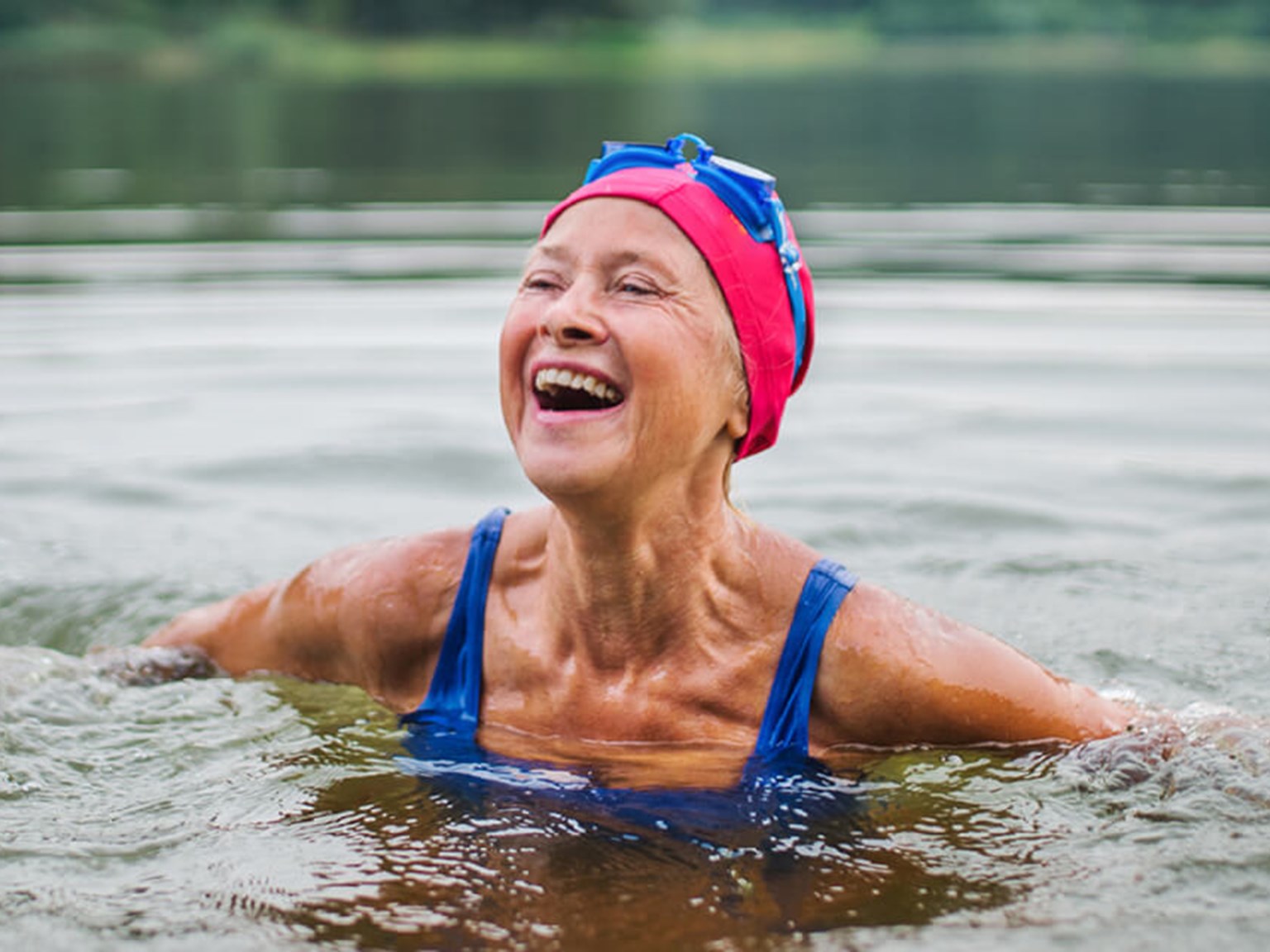 Happy older woman, wild swimming in a lake, wearing a pink swimming cap and blue goggles
