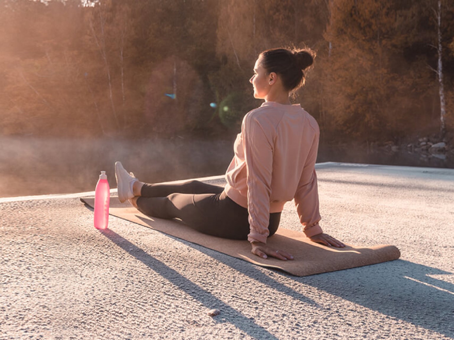 Woman in active wear, sat on a yoga mat next to a lake at sunrise.