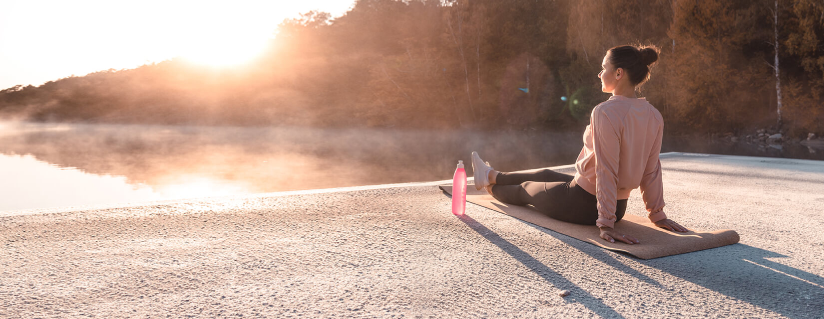 Woman in active wear, sat on a yoga mat next to a lake at sunrise.