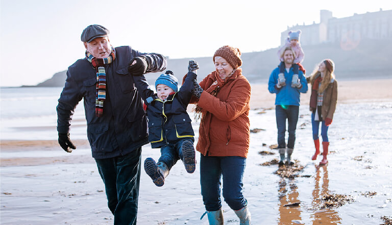 Grandparents swinging toddler by the arms on the beach whilst parents walk behind
