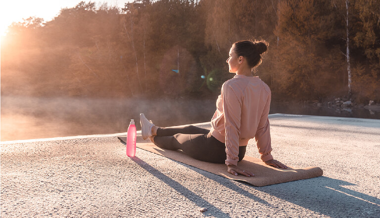 Woman sitting on a yoga mat on a dock, by a lake at dawn